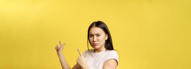 Covid-19 aşı konsepti. Hayal kırıklığına uğramış Asyalı kız reklam pankartına işaret ediyor, koronavirüsten aşı oluyor, sarı arka plan.