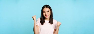Hevesli, sevimli Asyalı kız kazanıyor, harika haberlerle seviniyor, zafer dansı yapıyor, gülümsüyor, mutlu, mavi arka planda duruyor..