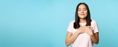 Gülümseyen Koreli kadın rüya görüyor, kalp ve kapalı gözlerle el ele tutuşuyor, mavi arka planda duran, ruhundaki anılara değer veriyor..