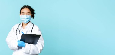 Covid-19 ve karantina konsepti. Gülümseyen Asyalı kadın doktor, maske takan hemşire elinde pano, hastanede nöbet tutuyor, mavi arka planda duruyor..