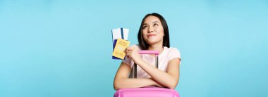 Seyahat hayalleri kuran mutlu Asyalı kadın turist, bavula yaslan, elinde covid-19 uluslararası aşı sertifikası, iki bilet ve pasaport, seyahat düşüncesi, mavi arka plan.