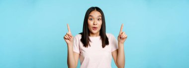 Şaşırmış Koreli kadın portresi huşu içinde, parmaklarını havaya kaldırarak, reklamını göstererek, mavi arka planda dikilerek,.
