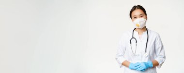 Solunum cihazında gülümseyen Asyalı kadın doktor, tıbbi maske ve lastik eldivenler, yardıma hazır, beyaz arka planda klinik üniforması içinde duruyor. Covid-19 ve karantina konsepti.