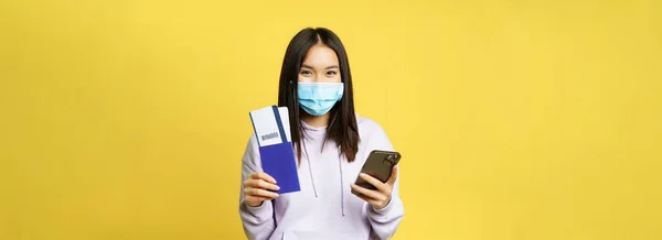 Asian tourist, girl in face mask, holding mobile phone, showing passport and flight tickets, booking hotel for vacation, going abroad, yellow background.