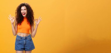 Cana yakın ve arkadaş canlısı beyaz bir kız. Kıvırcık saçlı, kısa şortlu. Onaylı jest yapıyor. Turuncu duvarın üstünde kameraya gülümseyen muhteşem partileri sevdiğini söylüyor..