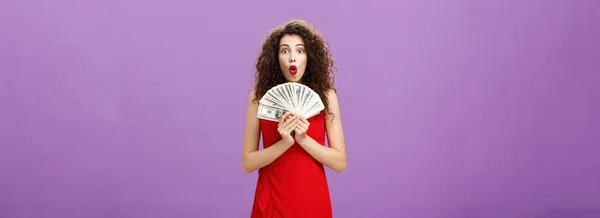 Studio shot of woman being lucky in casino winning. lots of cash holding money in hands saying wow with folded lips staring impressed and excited over purple wall in evening red dress.