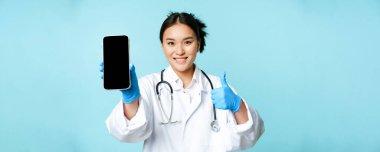 Web sağlık hizmeti, online yardım konsepti. Gülümseyen Asyalı kadın doktor ya da hemşire, akıllı telefon ekranı, uygulama arayüzü ve başparmaklar, tavsiye edilen web sitesi.