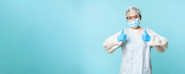 Optimistic asian nurse or doctor, wearing personal protective equipment, showing thumbs up, campaign from coronavirus prevention, blue background.