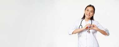 Gülümseyen Asyalı kadın doktor hastalarına bakıyor, kalp hareketi gösteriyor ve mutlu görünüyor, klinik üniforması içinde duruyor, beyaz bir geçmişi var..