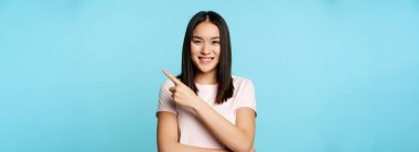 Gülümseyen genç Asyalı kadın 20 yaşında, sağ üst köşeyi işaret ediyor, reklam afişi gösteriyor, mavi arka plan.