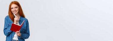 Yaratıcı, güzel, kızıl saçlı kız öğrenci, günlük kıyafetli genç, elinde kalem ve gülümseyen kamera, deftere notlar alıyor, günlük yazıyor, beyaz bir zemin hazırlıyor..