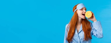 Lezzetli meyve sabah sağlıklı başlar. Belden yukarı portre çekici uykulu, uyku maskeli ve gecelikli güzel kızıl kız, portakal yiyen lezzetli, ayakta duran mavi arka plan.