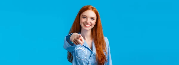 We need you. Cheerful and confident redhead girl making assured choice, pointing finger camera and smiling as decide, picking from variants, talking to person, standing in pyjama blue background.
