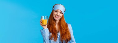 Günlük rutin, sabah ve sağlıklı yaşam tarzı konsepti. Çekici, enerjik, gecelikli bir kadın, uyku maskesi bir bardak meyve suyu kaldırıyor ve gülümsüyor, özenli vücut, mavi arka plan..