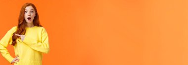 Dili tutulmuş ve etkilenmiş güzel, moda düşkünü, genç, modern, kızıl saçlı, düşük çeneli, nefes nefese kalmış, şaşırmış, kameraya bakan ve ilginç, harika bir ürünü işaret eden bir kız..