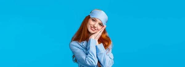 Have sweet dreams. Tender, lovely and feminine redhead teenage girl ready sleep tight, wearing sleeping mask and pyjama, lean on palms like pillow, smiling pleased and relaxed, blue background.