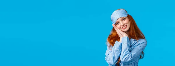 Have sweet dreams. Tender, lovely and feminine redhead teenage girl ready sleep tight, wearing sleeping mask and pyjama, lean on palms like pillow, smiling pleased and relaxed, blue background.
