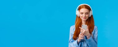 Belden yukarı portre kulaklıklı yakışıklı seksi üniversiteli kız, müzik dinliyor, sabahları haber yayını yapıyor, akıllı telefondan sosyal medyayı kontrol ediyor, pijamayla mavi arka planda duruyor..