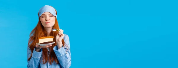 Delicious food, lifestyle and people concept. Delighted and happy cute redhead female in pyjama and sleep mask, close eyes and licking lips as eating tasty cake, holding spoon smiling.