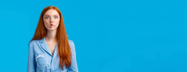 Silent like fish. Cute and funny young redhead female with long ginger hair, sucking lips as mimicking, fooling around in playful enthusiastic mood, wearing nightwear, standing blue background.