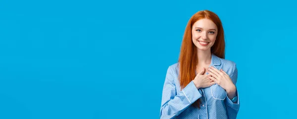 Feminine lovely redhead woman with long ginger hair, wearing pyjamas, hold hands on heart and smiling tenderly, having something precious hidden inside soul, standing blue background.