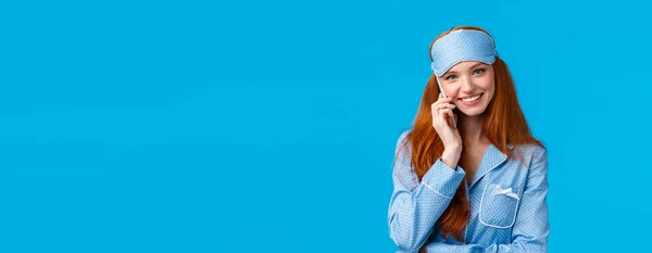 No you hang first. Lovely and beautiful redhead girlfriend talking phone till midnight, wearing sleep mask and nightwear, holding smartphone, calling boyfriend smiling silly, blue background.