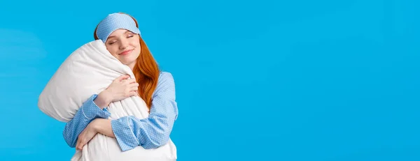Beauty, tenderness concept. Happy feminine cute redhead teenage girl in nightwear, sleep mask, close eyes and smiling delighted, finally went bed after long day, hugging pillow, blue background.