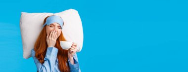 Uykucu, kızıl saçlı, uzun kızıl saçlı, uyku maskesi takan, gözleri kapalı, esneyen, sabah uyanan, yatakta kahve içen, yastıkta uzanan, mavi arka planda uzanan bir kız..