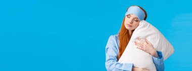 Güzellik, kadın ve yaşam tarzı konsepti. Rahat ve rahatlamış uyku maskesi ve pijama giymiş güzel kızıl saçlı kadın, uykulu gözler, tatlı rüyalar görüyor, yastığa sarılıyor, mavi arka planda dikiliyor..