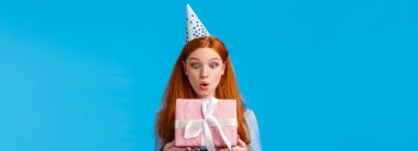 Vay canına. Etkilenmiş ve heyecanlı, çilli, kıvrılmış dudakları olan sevimli, çekici, kızıl saçlı kız. Alınan pembe paketli doğum günü hediyesine bak. Mutlu mavi arka plan..