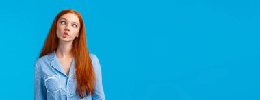 Sıra dışı ol. Kaygısız ve komik kızıl saçlı genç kız pijamayla pijama giyerken oda arkadaşına komik yüzler gösteriyor, gözlerini kısıyor ve balık yüzünü gösteriyor, mavi arka plan..