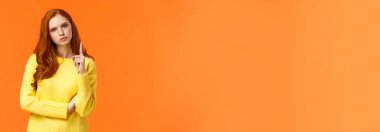 Kızıl saçlı kız, hayal kırıklığına uğramış kızgın bir yüze parmağını sallayarak, kaşlarını çatarak uyarıda bulunarak, kabul edilemez davranışları yasaklayarak, hoşnutsuz turuncu arka planda ayakta durarak onaylamadığını ifade ediyor..