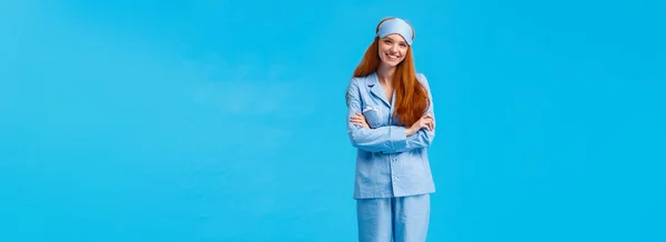 Vertical full-length shot happy cheerful caucasian foxy woman, teenage redhead girl in nightwear and sleep mask, standing blue background, cross arms over chest smiling. Copy space