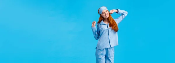 Full-length vertical shot cute and silly, teenage redhead girl, wearing lovely pyjama, waking up and stretching, had good sleep, smiling in morning, standing pleased blue background.