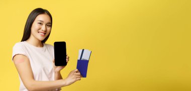 Gülümseyen Asyalı kız aşılandı, pasaport biletleri ve akıllı telefon ekranı, uygulama arayüzü gösteriliyor, sarı arka planda duruyor..