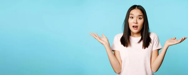 Confused asian girl spread empty hands sideways, shrugging shoulders clueless, standing in tshirt over blue background.