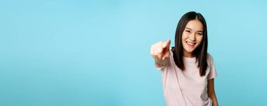 Sensin. Gülümseyen mutlu Asyalı kadın kamerayı işaret ediyor, insanları kutluyor, davet ediyor, mavi arka planda tişörtün üzerinde duruyor..