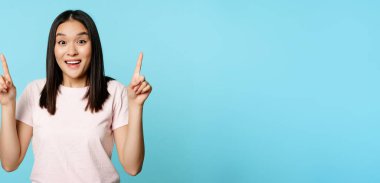 Hevesli Koreli kız reklamını yapıyor, işaret ediyor ve şaşırıyor, reklam anlaşmasını kontrol ediyor, mavi arka planda duruyor..