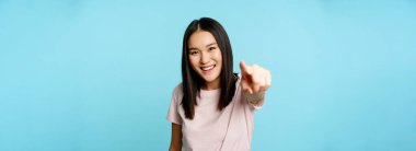 Sensin. Gülümseyen mutlu Asyalı kadın kamerayı işaret ediyor, insanları kutluyor, davet ediyor, mavi arka planda tişörtün üzerinde duruyor..