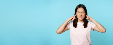 Sinirli Asyalı kız kulaklarını kapattı ve buruşturdu, yüksek sesle, sirenden rahatsız oldu, mavi arka planda tişört giydi. Boşluğu kopyala