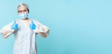 İyimser Asyalı hemşire ya da doktor, kişisel koruyucu ekipman giyiyor, baş parmaklarını gösteriyor, koronavirüs önleme kampanyası, mavi arka plan..