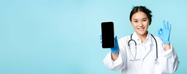 Web sağlık hizmeti, online yardım konsepti. Zeki telefon ekranı, uygulama arayüzü ve onaylı onay ve web sitesi önerisi gösteren kendinden emin Asyalı kadın doktor ya da hemşire..