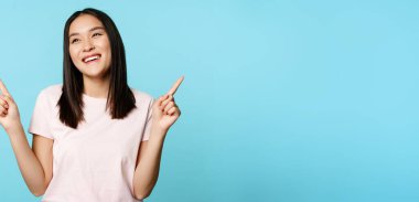 Gülümseyen esmer Asyalı kadın, iki tarafı da gösteriyor ve gülüyor, mağazayı seçiyor, mavi arka planda duruyor..