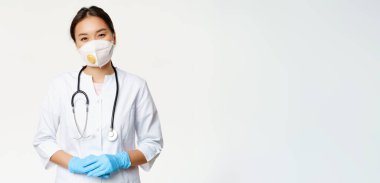 Solunum cihazında gülümseyen Asyalı kadın doktor, tıbbi maske ve lastik eldivenler, yardıma hazır, beyaz arka planda klinik üniforması içinde duruyor. Covid-19 ve karantina konsepti.