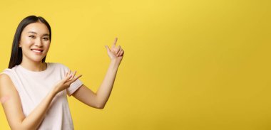 Covid-19 aşı konsepti. Omzunda yama olan gülümseyen Asyalı kız, sağlık afişini işaret ediyor, sarı arka plan.