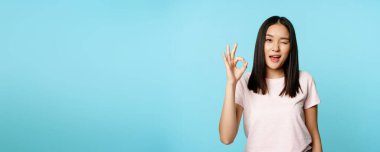 Tatmin olmuş Asyalı kız göz kırpıyor ve işaret veriyor. Onaylıyor, tavsiye ediyor, mavi arka plan üzerinde tişört giyiyor..