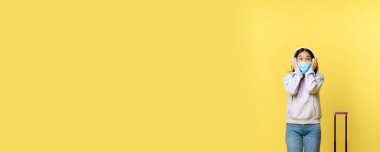 Mutlu, gülümseyen Asyalı turist, kulaklıklı kız, havaalanında müzik dinleyen, tıbbi maske takan, bavulla ayakta duran, sarı arka plan.