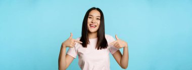 Kendine güvenen Koreli kadın gülümsüyor, kendini beğenmiş bir ifade takınıyor, kendi reklamını yapıyor, gönüllü oluyor, kazanan oluyor, mavi arka planda duruyor..