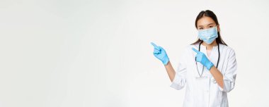 Asyalı kadın sağlık görevlisinin portresi solu işaret ediyor, yüz maskesi ve lastik eldiven takıyor, beyaz arka planda klinik üniforması içinde duruyor..
