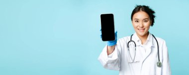 Web sağlık hizmeti, online yardım konsepti. Gülümseyen Asyalı kadın doktor, akıllı telefon uygulaması arayüzü, mobil ekran, mavi arka planda üniforma içinde duruyor..